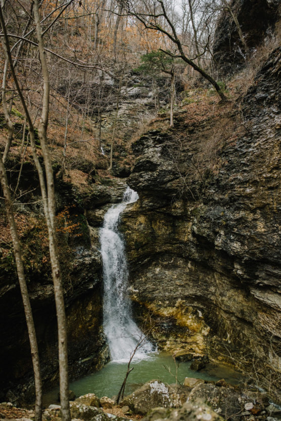A small waterfall flows over layered rocks into a shallow green pool, surrounded by bare trees and rugged cliffs in a forest during late autumn or early winter. by Justin Salem Meyer