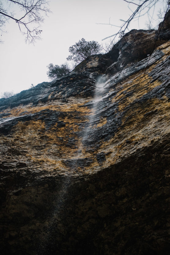 A thin waterfall trickles down a rugged, rocky cliff face with sparse vegetation, viewed from below against a pale, overcast sky. by Justin Salem Meyer