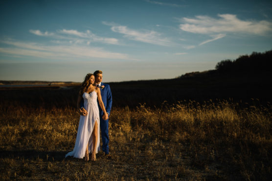 A couple in formal attire stands together in a sunlit field at sunset. The woman wears a flowing white dress, and the man wears a blue suit. They gaze into the distance, surrounded by tall grass under a dramatic sky. by Justin Salem Meyer