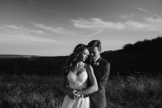 A black and white photo of a bride and groom embracing outdoors in a field. The bride is wearing a lace dress and flower crown, while the groom hugs her from behind, both looking peaceful and happy under a partly cloudy sky. by Justin Salem Meyer
