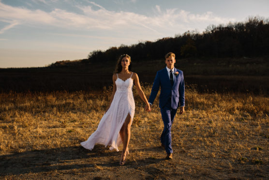 A bride in a white dress and a groom in a blue suit walk hand-in-hand across a sunlit, grassy field, with trees and a blue sky in the background. by Justin Salem Meyer