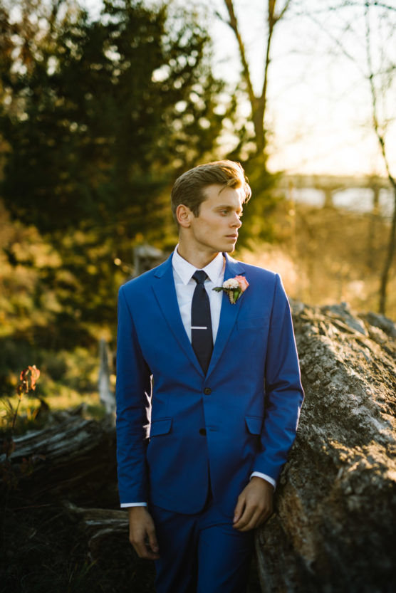 A man in a bright blue suit with a boutonniere stands outdoors by a fallen tree, bathed in warm, golden sunlight. The background features trees and soft, natural foliage. by Justin Salem Meyer