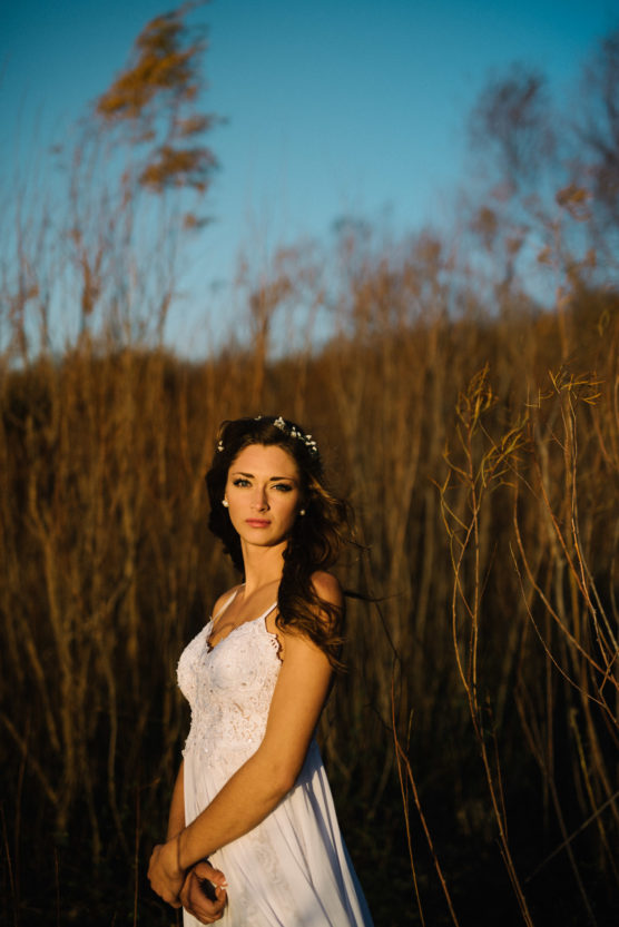 A woman in a white dress with a floral headband stands in a sunlit field of tall dry grass, looking at the camera, with soft evening light and a clear blue sky behind her. by Justin Salem Meyer