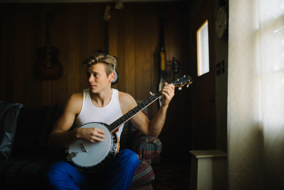 A young man in a white tank top and blue pants sits on a plaid couch playing a banjo in a warmly lit, wood-paneled room with guitars hanging on the wall behind him. by Justin Salem Meyer
