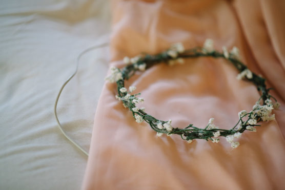 A delicate floral crown with small white flowers and green stems rests on a soft pink fabric, partially draped over a white surface. by Justin Salem Meyer