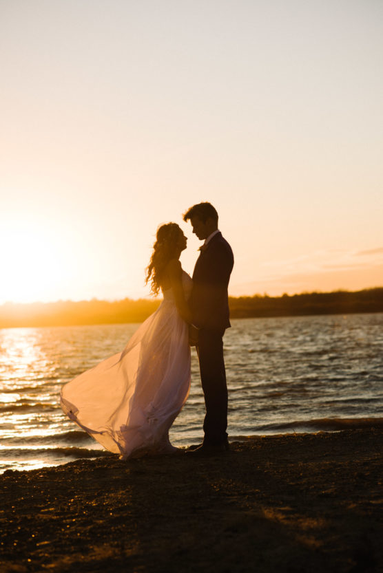 A couple stands close together on a beach at sunset, silhouetted against the glowing sky. The woman wears a flowing dress that catches the light breeze, while the man stands facing her near the water’s edge. by Justin Salem Meyer
