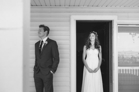 A bride in a white dress and a groom in a suit stand near a doorway. The groom faces left, smiling, while the bride stands in the doorway, looking at him and smiling. The photo is in black and white. by Justin Salem Meyer