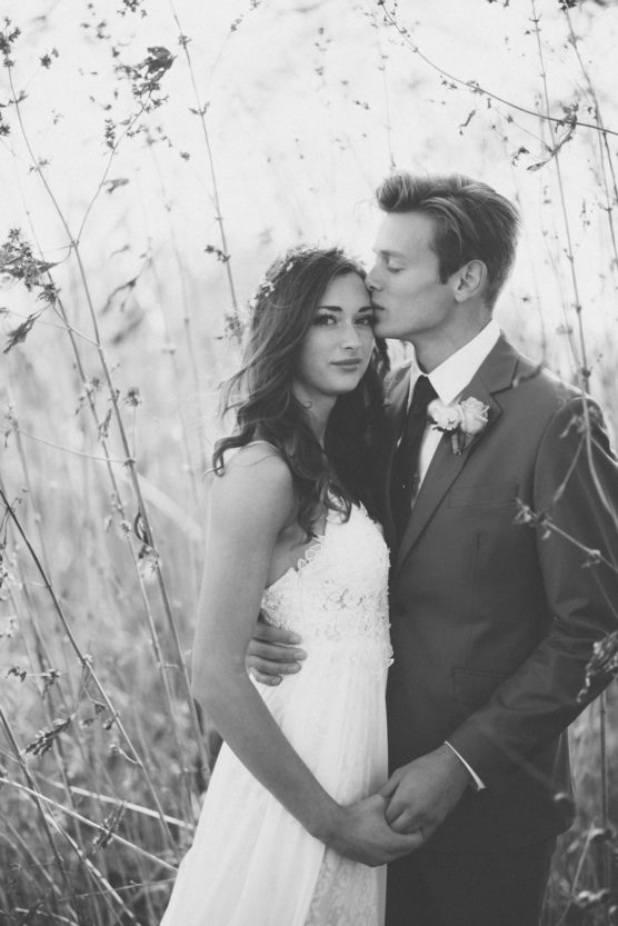 A bride and groom stand close together in a field of tall grass. The groom kisses the bride’s forehead while holding her waist and hand. Both wear formal attire; the scene is in black and white. by Justin Salem Meyer