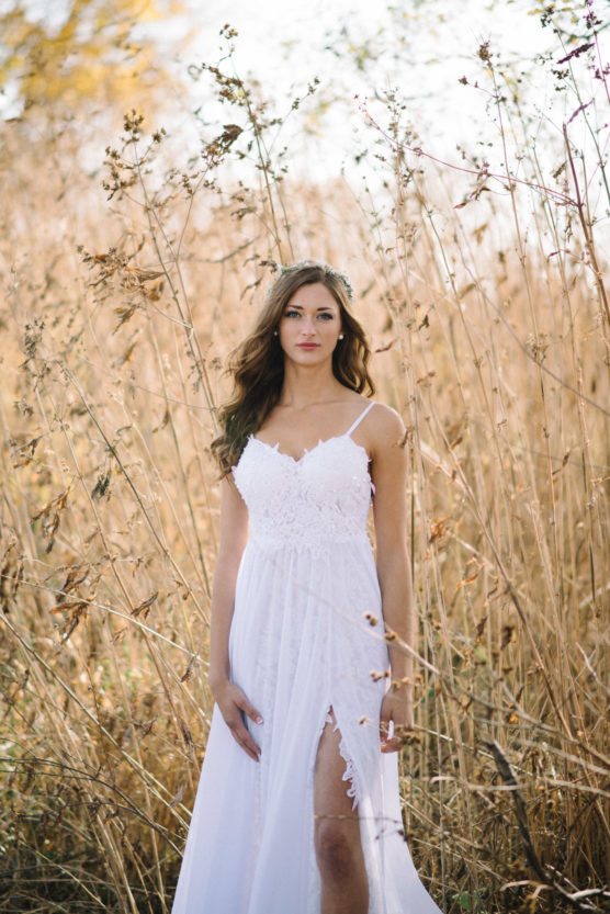 A woman in a white, sleeveless lace dress stands in a field of tall, dry grass, looking at the camera with a calm expression. The sunlight creates a warm, natural atmosphere. by Justin Salem Meyer