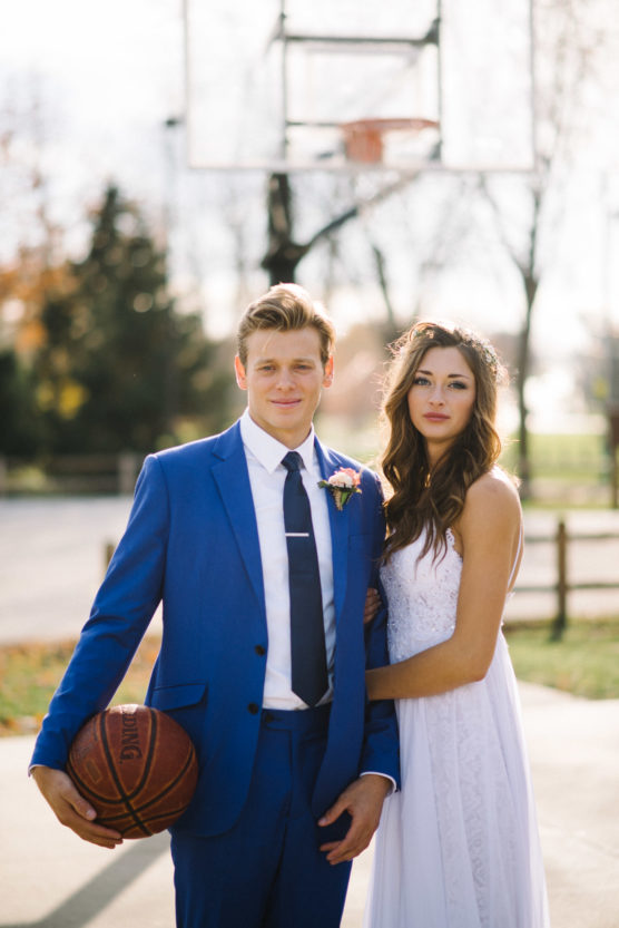 A groom in a blue suit holding a basketball stands beside his bride in a white dress. They pose outdoors on a basketball court with a hoop and trees in the background. by Justin Salem Meyer