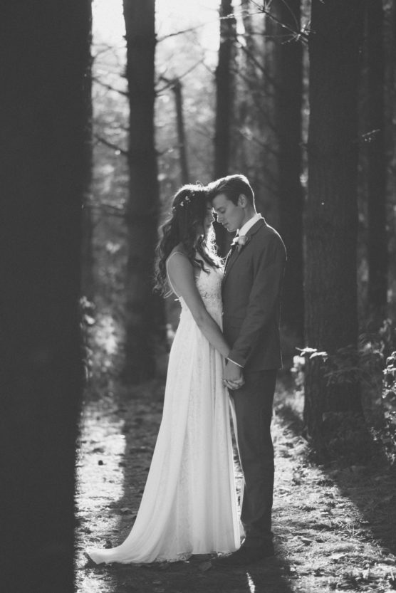 A bride and groom stand in a sunlit forest, holding hands and touching foreheads, sharing an intimate moment. The image is in black and white. by Justin Salem Meyer