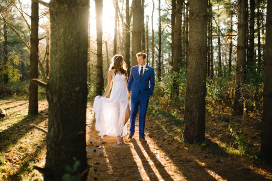 A couple dressed formally, the woman in a white dress and the man in a blue suit, walk hand-in-hand along a sunlit forest path, with light streaming through tall trees around them. by Justin Salem Meyer