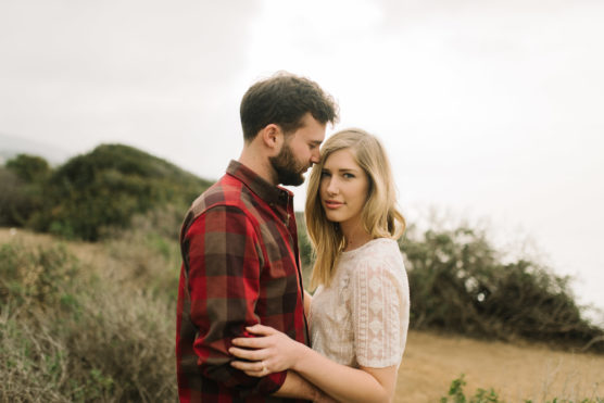 A couple stands close together outdoors; the man in a red plaid shirt rests his forehead on the woman’s, who wears a white lace top and looks at the camera. Greenery and an overcast sky are visible in the background. by Justin Salem Meyer