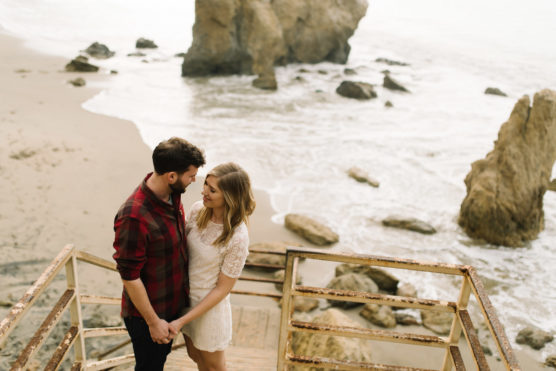 A couple stands on rusted stairs by a rocky beach, holding hands and gazing at each other, with the ocean and large rocks visible in the background. by Justin Salem Meyer