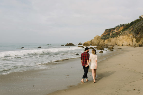 A couple walks barefoot on a sandy beach near the shoreline, holding hands. Rocky cliffs and gentle waves are visible in the background under a cloudy sky. by Justin Salem Meyer