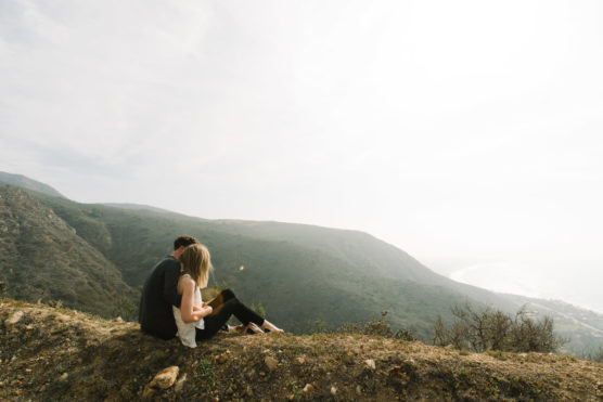 A couple sits on a grassy hillside overlooking a foggy, expansive landscape of rolling green hills and a distant coastline under a cloudy sky. by Justin Salem Meyer