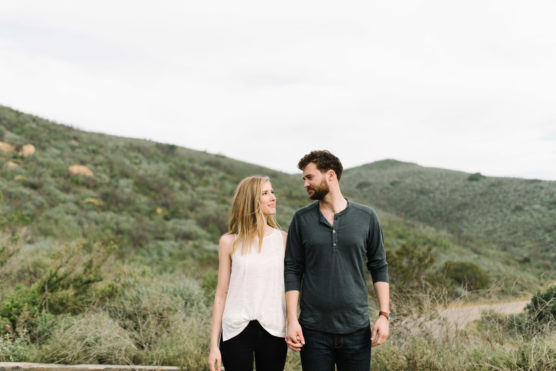 A man and woman hold hands and walk outdoors, surrounded by green hills and vegetation under an overcast sky. They look at each other and smile, appearing relaxed and happy. by Justin Salem Meyer