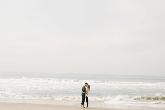 A couple stands close together and kisses on a sandy beach, with gentle waves and a cloudy sky in the background. The scene feels calm and romantic with wide open space around them. by Justin Salem Meyer