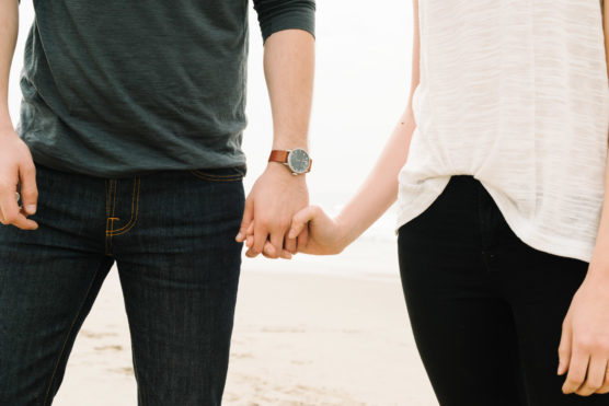 A cropped image of two people holding hands on a beach. One person wears a dark long-sleeve shirt, blue jeans, and a watch; the other wears a white short-sleeve top and black jeans. Sand is visible in the background. by Justin Salem Meyer