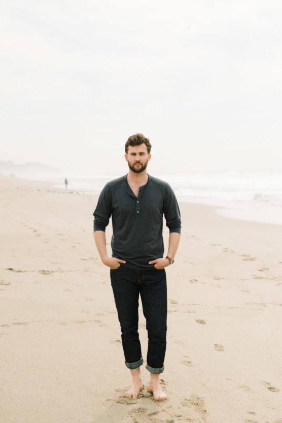 A man with a beard stands barefoot on a sandy beach, wearing a dark long-sleeve shirt and rolled-up jeans. He has his hands in his pockets, with waves and a cloudy sky in the background. by Justin Salem Meyer