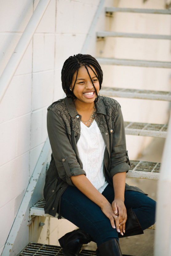 A young woman with braided hair sits on metal stairs, smiling. She wears a green jacket over a white shirt, jeans, and black boots. The background features a white wall and railings. by Justin Salem Meyer