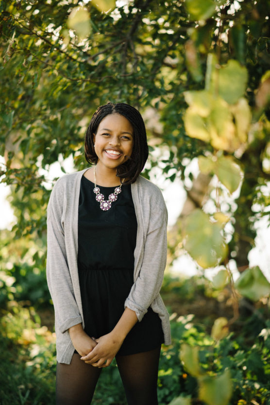 A young woman stands outdoors, smiling, wearing a black dress, light gray cardigan, and statement necklace. She is surrounded by green foliage and sunlight filters through the trees. by Justin Salem Meyer