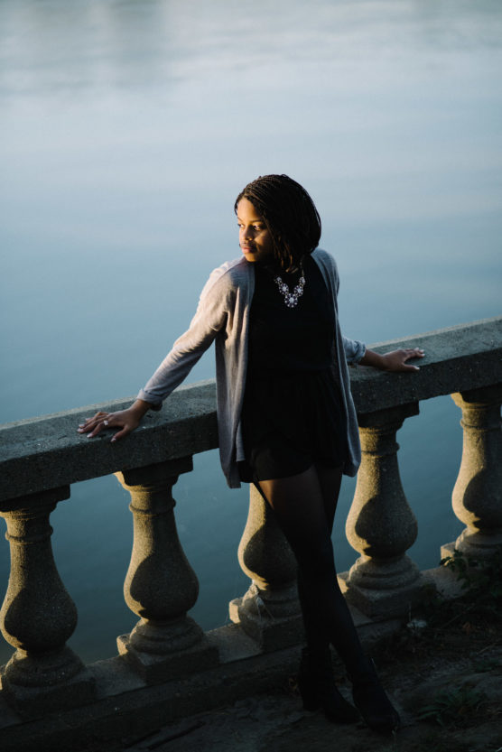 A woman in a black outfit and gray cardigan stands by a stone balustrade beside a body of water, looking to her left. She is illuminated by soft, natural light. by Justin Salem Meyer