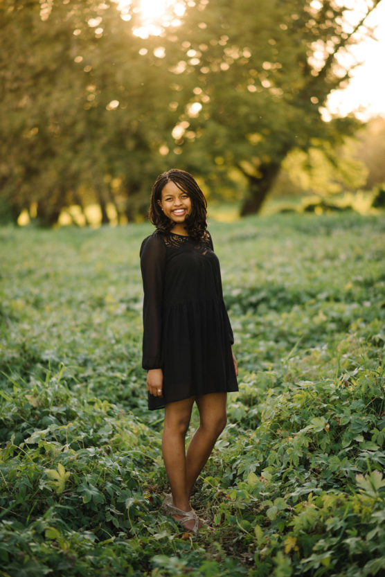 A young woman in a black dress stands smiling in a green field with tall trees and sunlight filtering through the leaves in the background. by Justin Salem Meyer