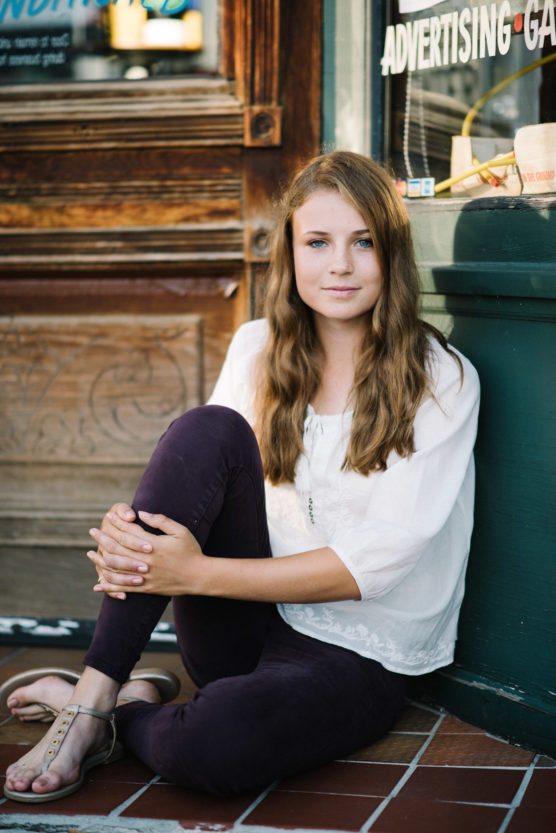 A young woman with long brown hair, wearing a white blouse, dark pants, and sandals, sits on the ground with her knees up, leaning against a storefront window and looking at the camera with a slight smile. by Justin Salem Meyer
