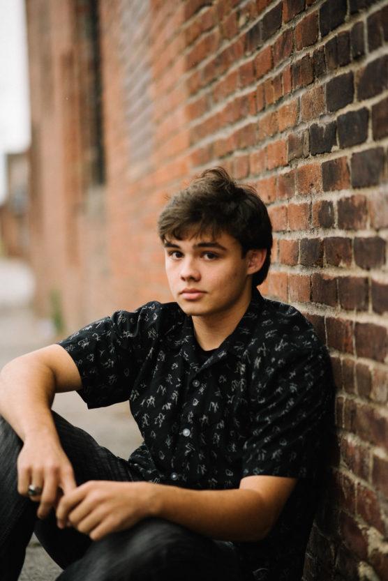 A young person with short brown hair sits against a brick wall, wearing a black patterned shirt and looking at the camera with a neutral expression. The background is blurred, focusing on the subject. by Justin Salem Meyer