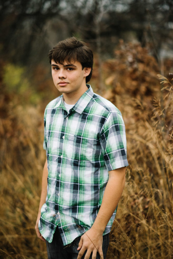 A young person with short brown hair wearing a green and white plaid shirt stands outdoors in a field of tall, dry grass, looking at the camera with a neutral expression. The background is blurred with autumn colors. by Justin Salem Meyer