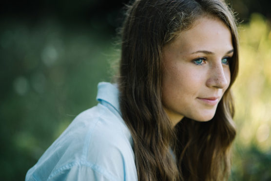A young woman with long brown hair and blue eyes, wearing a light blue shirt, gazes thoughtfully to the right. She is outdoors with a blurred green background. by Justin Salem Meyer