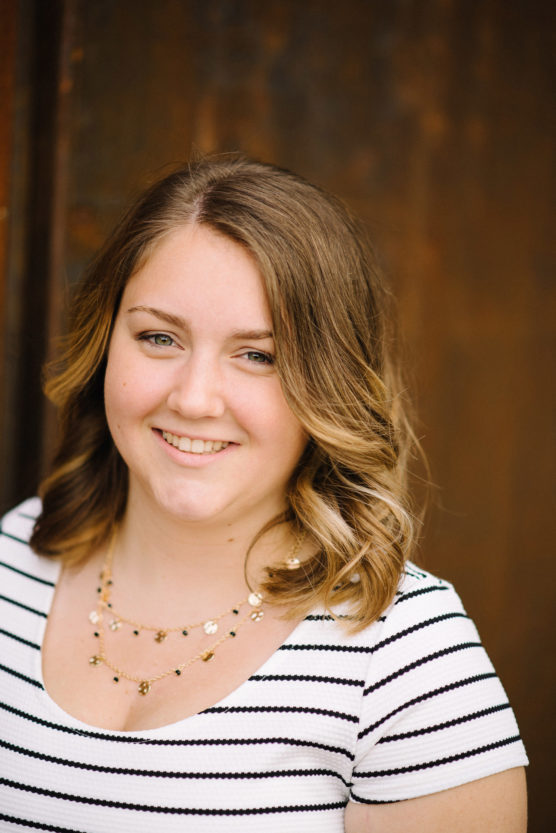 A woman with shoulder-length wavy blonde hair, wearing a white shirt with black stripes and a gold necklace, smiles in front of a blurred brown background. by Justin Salem Meyer