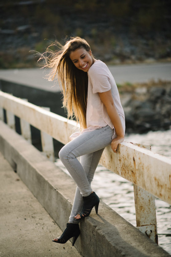 A young woman with long hair sits on a rusty guardrail by the water, smiling with her eyes closed. She wears a light pink t-shirt, light gray jeans, and black heeled sandals. Sunlight highlights her hair. by Justin Salem Meyer