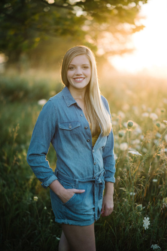 A young woman with long blonde hair, wearing a denim romper, stands smiling in a sunlit field with tall grass and wildflowers, with the warm glow of the sunset behind her. by Justin Salem Meyer