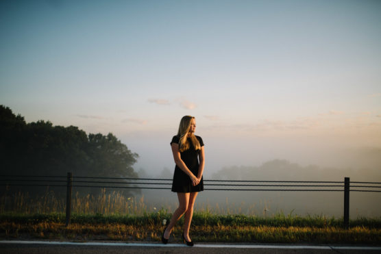 A woman in a black dress stands by a roadside guardrail at sunrise, with misty trees and a soft, hazy sky in the background. by Justin Salem Meyer