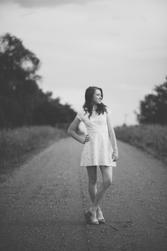 A woman in a short, light dress stands on a deserted country road, surrounded by trees and fields, looking to the side. The image is in black and white. by Justin Salem Meyer