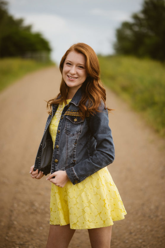 A young woman with long red hair smiles at the camera while standing on a rural dirt road. She is wearing a yellow dress with a floral pattern and a blue denim jacket. Green trees and grass line the road in the background. by Justin Salem Meyer