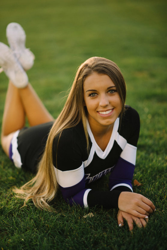 A young woman in a black and white cheerleading uniform lies on her stomach on green grass, smiling at the camera with her legs bent and feet crossed in the air behind her. by Justin Salem Meyer