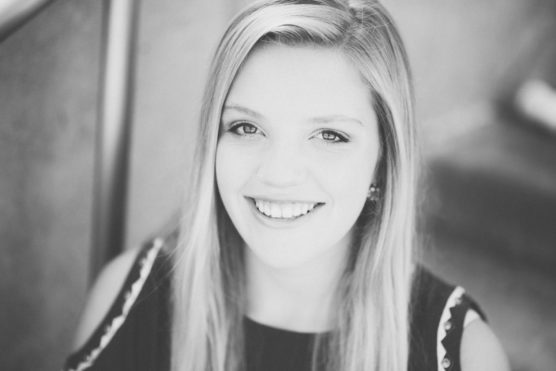 Black and white portrait of a young woman with long, straight hair, smiling at the camera. She is wearing a sleeveless top with patterned details on the shoulders. The background is softly blurred. by Justin Salem Meyer