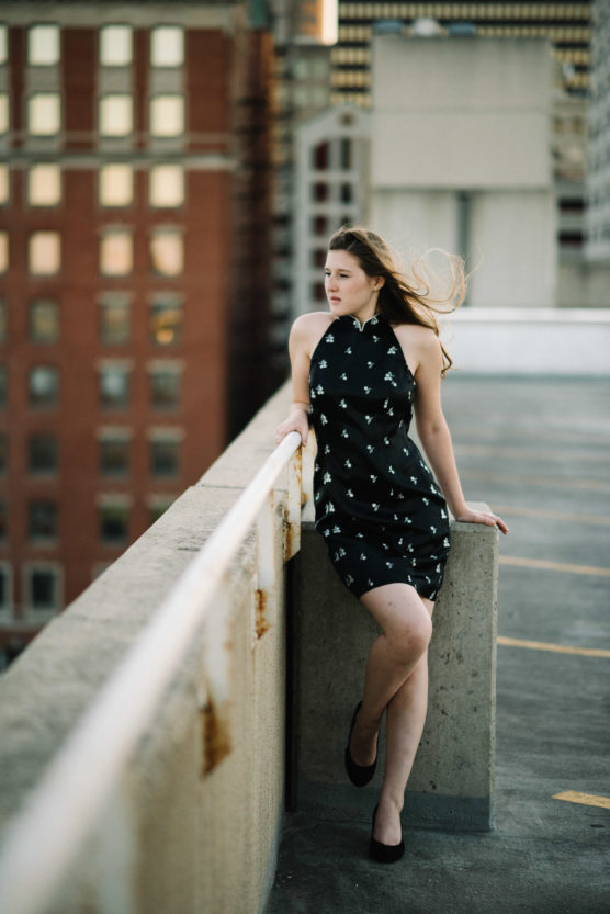 A woman in a black dress with white patterns stands on a rooftop parking lot, leaning against a concrete barrier, with city buildings in the blurred background and wind blowing her hair. by Justin Salem Meyer