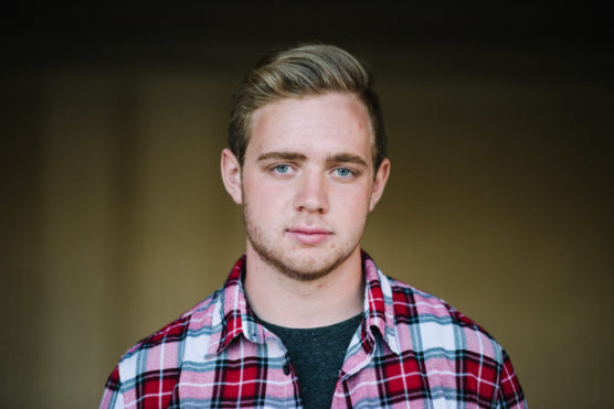 A young man with short blond hair and blue eyes, wearing a red and white plaid shirt over a dark T-shirt, looks directly at the camera with a neutral expression. The background is softly blurred. by Justin Salem Meyer