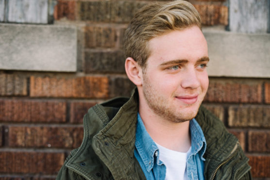 A young man with short blond hair and a light beard stands in front of a brick wall, wearing a green jacket over a blue denim shirt and white t-shirt, looking slightly to the right with a slight smile. by Justin Salem Meyer