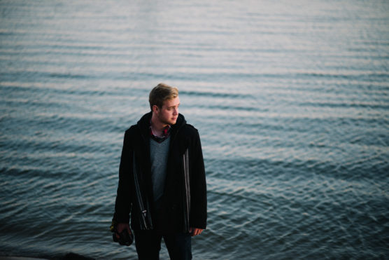 A young man in a dark coat stands on the shore, holding a camera by his side, looking to his left. The calm water with gentle ripples stretches out behind him under soft, natural light. by Justin Salem Meyer