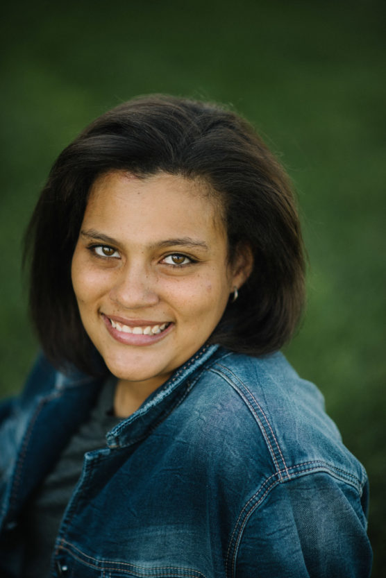 A person with medium-length dark hair, wearing a denim jacket, smiles while looking up at the camera against a blurred green outdoor background. by Justin Salem Meyer