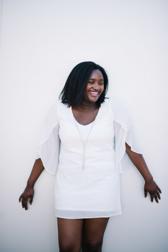 A woman with shoulder-length dark hair, wearing a white dress and a long necklace, stands against a plain white background, smiling and looking to the side with her arms relaxed by her sides. by Justin Salem Meyer
