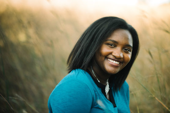 A young woman with straight black hair and a blue top smiles warmly at the camera while sitting outdoors in a field of tall grass. by Justin Salem Meyer