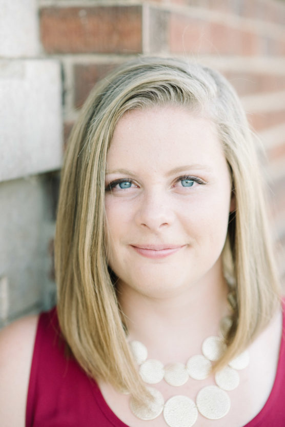 A woman with straight blonde hair, blue eyes, and a slight smile stands in front of a brick wall. She is wearing a sleeveless maroon top and a chunky circular white necklace. by Justin Salem Meyer
