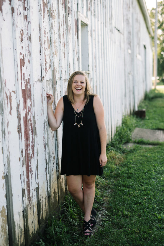 A woman in a sleeveless black dress and sandals stands smiling against a worn, white wooden wall outside, with grass and greenery surrounding her. by Justin Salem Meyer