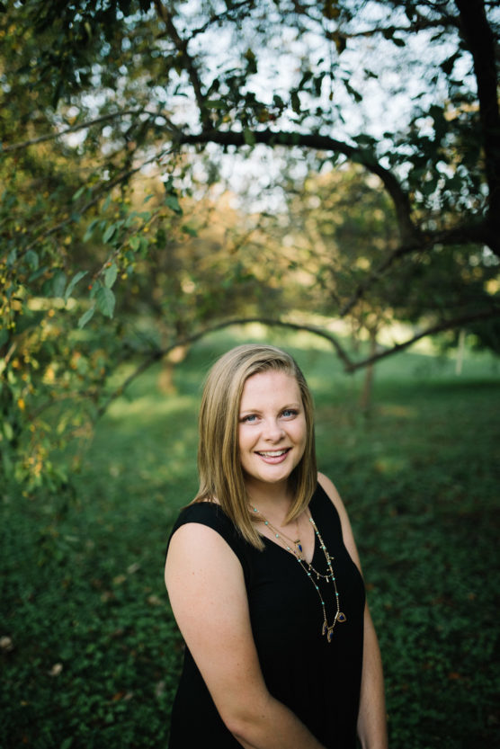 A young woman with straight blonde hair, wearing a sleeveless black dress and a long necklace, stands outside smiling with green trees and grass in the background. by Justin Salem Meyer